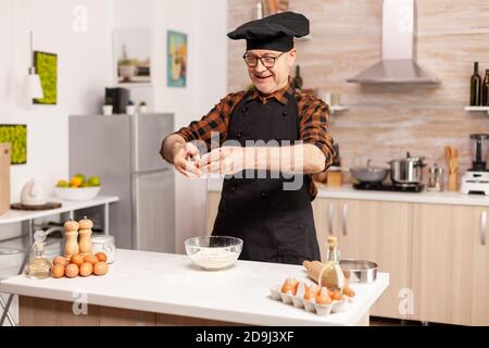 Felice uomo anziano che cricchiola le uova sulla farina di grano mentre prepara la ricetta tradizionale. Cuoco di pasta anziano che coglie l'uovo sulla ciotola di vetro per la ricetta della torta in cucina, mescolando a mano, impastando Foto Stock