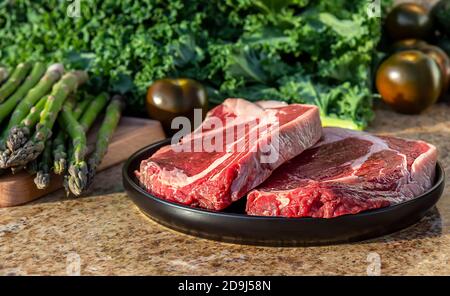 Carne cruda, bistecca di manzo su piatto nero con kale, asparagi, broccolini e pomodori sullo sfondo. Foto Stock