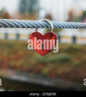 Lucchetto a cuore rosso sul cavo in acciaio della recinzione del ponte nella città di Kharkov in Ucraina. Paesaggio urbano non focalizzato in colori autunnali sullo sfondo. Messa a fuoco selettiva. Copia Foto Stock