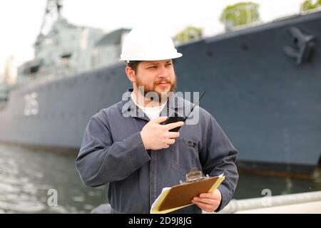 Capitano europeo marino che parla da walkie talkie radio e in piedi vicino nave con documenti in mani. Foto Stock