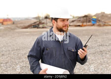 Ritratto di caposquadra in piedi sul cantiere, parlando da VHF walkie talkie e tenere progetti. Foto Stock