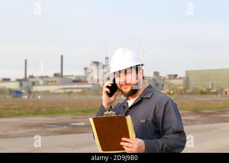 Ritratto di caucasio caucasiano in piedi sul luogo di costruzione, parlando da walkie talkie e tenendo carte. Foto Stock