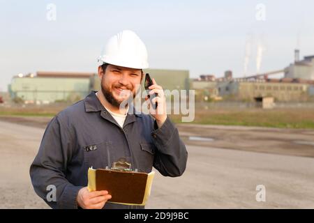 Ritratto di caposquadra in piedi sul sito di costruzione, parlando da walkie talkie e tenendo carte. Foto Stock