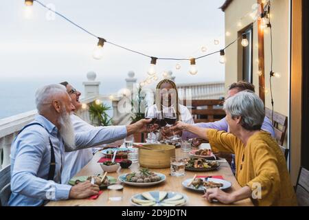 Felici anziani multirazziali che tostano insieme con bicchieri di vino rosso Cena nel patio della casa - concetto di gente anziana di stile di vita Foto Stock