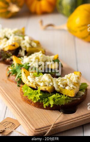 Panini tostati con formaggio brie, zucca al forno e insalata, rucola e mozzarella, deliziosi spuntini vegetali su tavola di legno Foto Stock