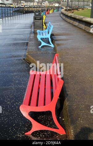 Largs, Ayrshire, Scozia, Regno Unito. Lungomare, con panchine colorate. Una fila di panchine di parco dipinte con colori luminosi lungo il lungomare della località balneare Ayrshire. Contenitori per rifiuti distanziati tra i banchi Foto Stock