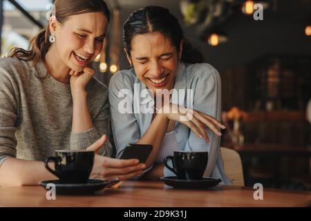 Due amici di sesso femminile seduti in una caffetteria e sorridenti. Due donne attraenti che usano un cellulare mentre si rilassano insieme in un caffè. Foto Stock