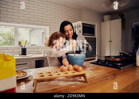 Madre sorridente che aiuta la figlia a mescolare le uova e versare l'acqua per impasto in cucina Foto Stock