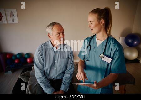 Sorridente giovane medico che tiene una tavoletta digitale che spiega la procedura di trattamento a. paziente anziano Foto Stock