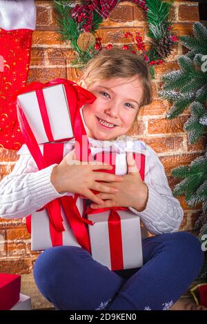 La ragazza bionda caucasica piccola cute apre le scatole dei regali di Natale. Home sfondo con abete, regali di Natale, decorazioni. Capodanno e Natale Foto Stock