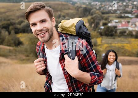 Turista sorridente a macchina fotografica mentre cammina vicino afroamericana donna su sfondo sfocato all'aperto Foto Stock