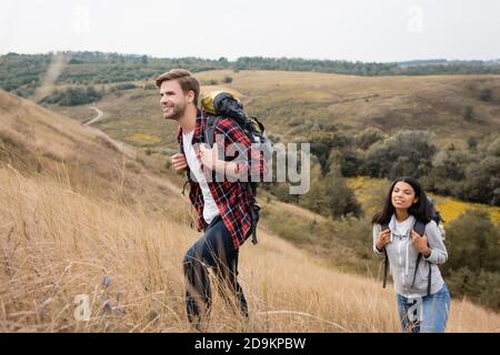 Uomo sorridente con zaino che cammina sulla collina vicino afroamericano ragazza durante la vacanza Foto Stock