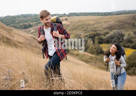 Uomo sorridente con zaino che cammina vicino afroamericana fidanzata sopra collina con erba Foto Stock
