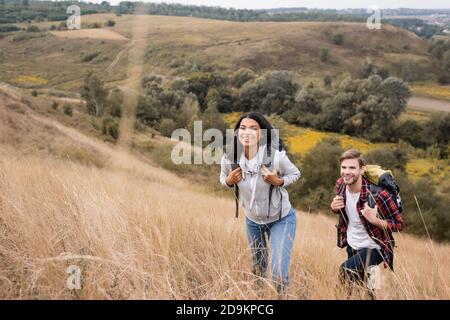 Coppia interracial con zaini che camminano su collina con erba durante viaggio Foto Stock