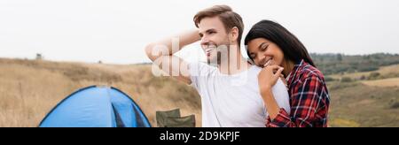 Donna afroamericana sorridente mentre abbracciando il ragazzo con tenda su sfondo sfocato su prato, banner Foto Stock