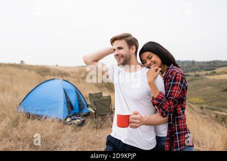 Donna afroamericana con gli occhi chiusi che abbracciano il ragazzo con la tazza vicino alla tenda sul primo piano sfocato Foto Stock