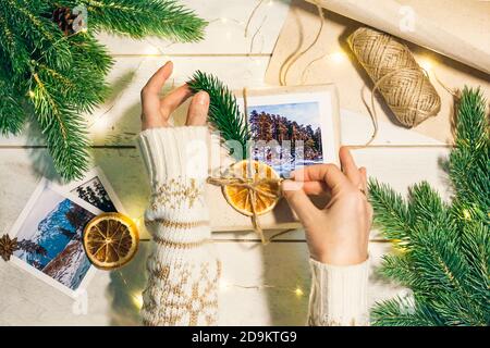 Mani donne che tengono regalo di natale imballato con le foto delle montagne in stile eco-friendly: Carta ruvida naturale, corda di iuta, ramoscelli di abete rosso, sci vintage Foto Stock