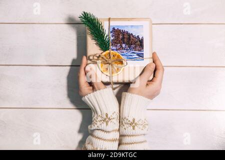 Mani donne che tengono regalo di natale imballato con foto delle montagne in stile eco-friendly su sfondo di legno. Viaggia come regalo. Foto Stock