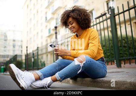 Ritratto integrale giovane donna afroamericana seduta per strada utilizzando il cellulare Foto Stock