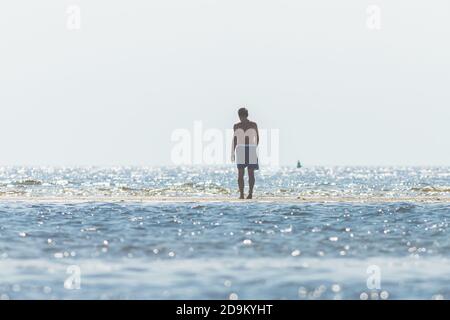 Beach Recreation - l'uomo in tronchi da bagno si affaccia sull'acqua e guarda le onde. Foto Stock