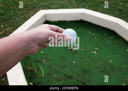 Mano bianca che tiene la sfera di golf sopra il relativo foro al campo. Giocare a mini golf su erba verde artificiale all'aperto. Definizione di obiettivi e concetto vincente. Divertimento. Foto Stock