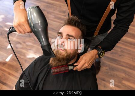 Il ragazzo caucasico con la barba è pettinato da un barbiere ispanico nel barbiere con un pettine e un asciugacapelli. Aereo zenithal. Foto Stock