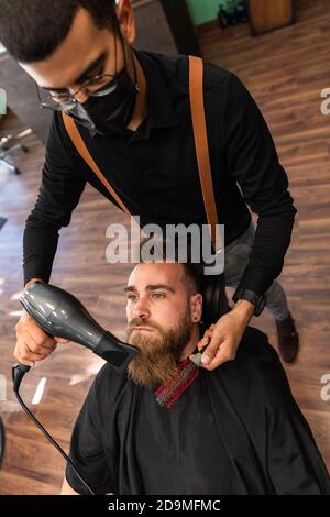 Il ragazzo caucasico con la barba è pettinato da un ispanico barbiere nel barbiere con pettine e asciugacapelli e maschera nera per coronavirus pandemic protec Foto Stock