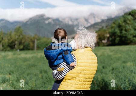 Vista posteriore di una nonna che indossa una maschera medica che porta suo nipote in un campo Foto Stock