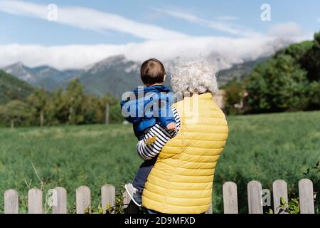 Vista posteriore di una nonna che indossa una maschera medica che porta suo nipote in un campo Foto Stock