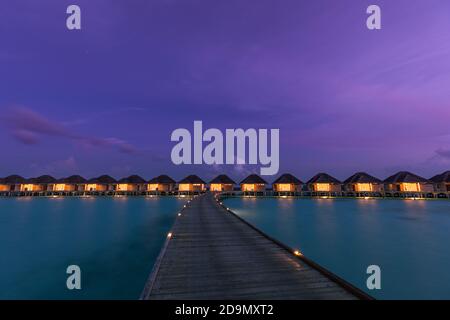 Bungalow sull'acqua nelle isole Maldive. Paradiso tropicale isola destinazione, luci LED con lungo molo sotto il cielo notturno crepuscolo. Paesaggio esotico Foto Stock