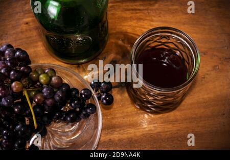 Piatto di un bicchiere di vino su un tavolo di legno, accanto ad una ciotola di cristallo piena di uva matura, e una bottiglia verde Foto Stock