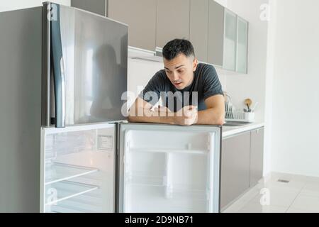 Il ragazzo è triste vicino al frigorifero vuoto senza cibo. Pubblicità del servizio di consegna del cibo. Foto Stock