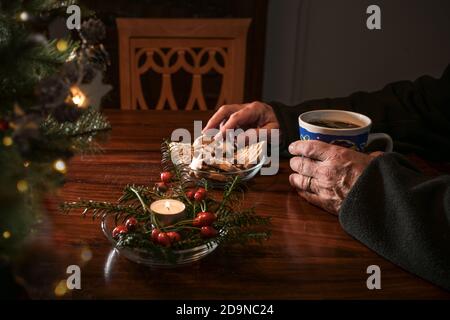 Mani di un uomo anziano seduto da solo ad un tavolo con biscotti di Natale, caffè e decorazione festosa accanto ad una sedia vuota, vacanze solitarie Foto Stock