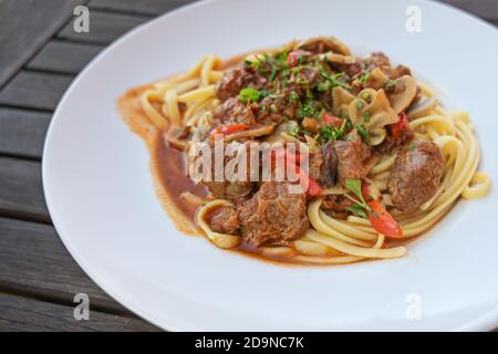 Ragù di manzo, peperoni rossi e funghi con contorno di prezzemolo sulla pasta in un piatto bianco su un tavolo di legno rustico scuro, spazio copia, selezionato fo Foto Stock