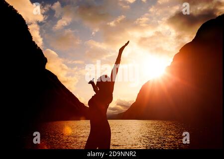 Silhouette di una donna con le braccia allungate all'alba da un lago Foto Stock