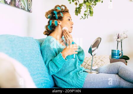 Bella giovane donna nel trattamento di cura della pelle a casa seduta sul divano - blu e bianco colori e bella donne con arricciacapelli e acqua tonica sul viso - concetto di attività di bellezza Foto Stock