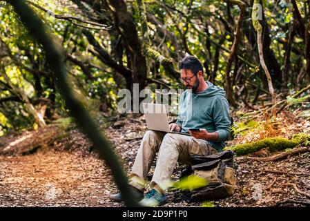 L'uomo adulto lavora con il computer portatile e la connessione telefonica in roaming nel mezzo della foresta verde selvaggia - concetto di nomade digitale e moderni lavoratori intelligenti ovunque internet connesso Foto Stock