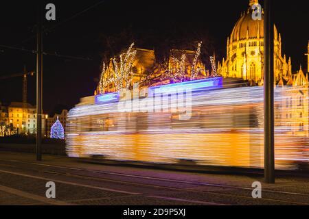 Tram chiaro decorato in modo festivo, Fenyvillamos, in movimento con il Parlamento di Ungheria a piazza Kossuth di notte. Stagione natalizia a Budapest, Ungheria Foto Stock