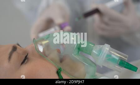 Una paziente donna sta ricevendo ventilazione, ossigeno perché è infettata dal coronavirus. Un medico sta mostrando una provetta positiva sullo sfondo. Foto Stock