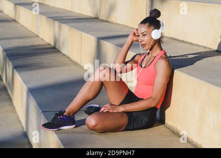 Ritratto orizzontale di attraente giovane afroamericana vestita sportswear e le moderne cuffie bianche che si riposano su alcuni fasi Foto Stock