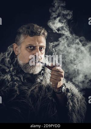 Uomo dal bearded solido in uno studio di pipe per fumatori con mantle di pelliccia ritratto su sfondo nero Foto Stock