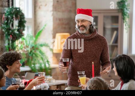 Felice uomo maturo nel cappello di Santa facendo toast durante la famiglia cena al tavolo delle feste Foto Stock