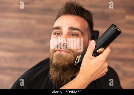 Vista dall'alto di un cliente in un barbiere, la sua barba viene tagliata e tagliata con un rasoio elettrico Foto Stock