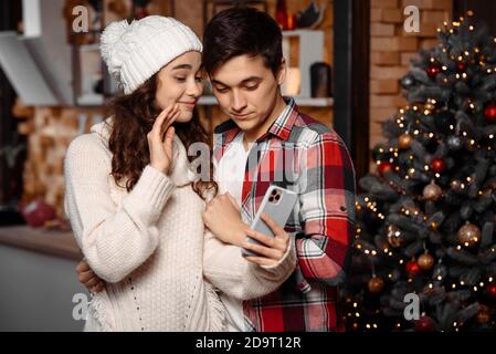 Felice giovane coppia comunica via video su smartphone, in piedi vicino decorato albero di natale godendo di trascorrere il tempo insieme. Momenti accoglienti e gioiosi nelle vacanze invernali. Concetto di pubblicità Foto Stock