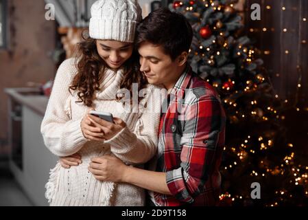 Felice giovane coppia che controlla i messaggi di Natale su uno smartphone abbracciando in piedi vicino decorato albero di natale godendo di passare il tempo insieme. Momenti accoglienti e gioiosi nelle vacanze invernali. Concetto di pubblicità Foto Stock