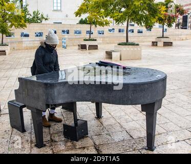 Gerusalemme, Israele - 5 novembre 2020: Una giovane donna che indossa una maschera protettiva, suona un pianoforte di strada a Gerusalemme, Israele, in un giorno piovoso. Foto Stock