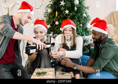 Gruppo di giovani amici seduti accanto ad un albero di Natale ben decorato, scambiando regali di Natale e bevinkink chapmagne. Foto Stock