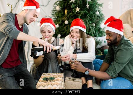 Gruppo di giovani amici seduti accanto ad un albero di Natale ben decorato, scambiando regali di Natale e bevinkink chapmagne. Foto Stock