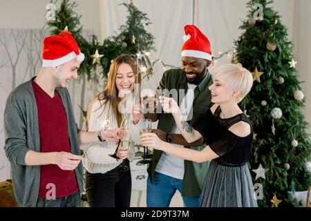 Uomini felici e donne soddisfatte in cappelli di Santa bere bicchieri di champagne mentre comunicare e ridere di festa di Capodanno. Tengono il bengala Foto Stock