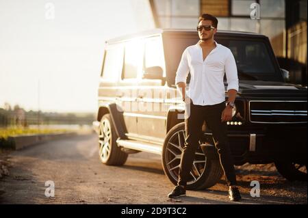 Uomo asiatico solido in camicia bianca e occhiali da sole posati vicino nero mafia suv auto. Foto Stock